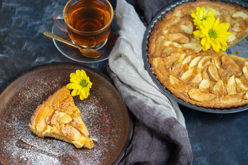 Charlotte apple pie in a round shape, decorated with fan-laced apple slices from the top, a piece of cake next to it, a cup of tea, autumn leaves, all against a dark background. 