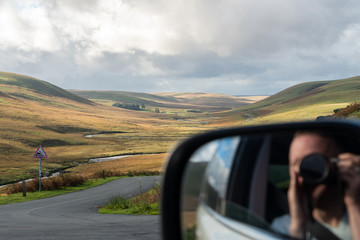 Reflection of photographer in car wing mirror looking onto valleys with shadows from clouds