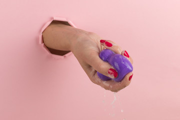 Girl hand holds a beauty blender for makeup from a hole in a pink background, copy space. Sponge in soapy foam, how to clean.