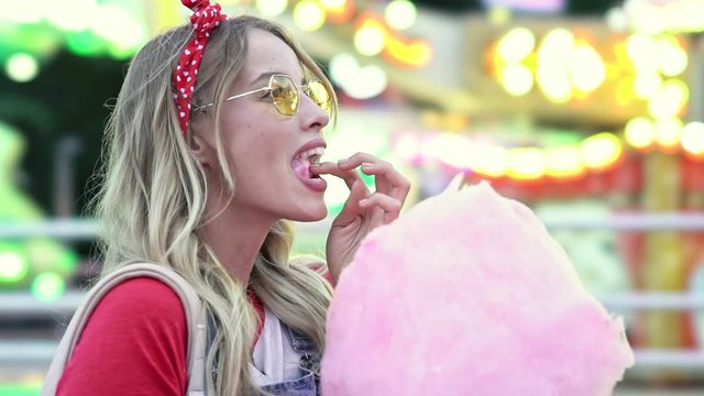 Side view of happy young blonde woman laughing and enjoying eating cotton candy while spending time at amusement park