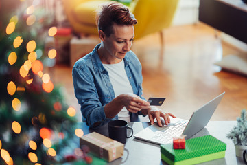 Woman sitting by the Christmas tree using laptop and paying online with credit card