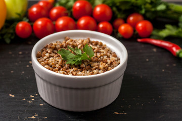 Cooked buckwheat porridge in a deep plate on a black wooden background with vegetables.
