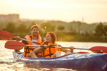 Happy young caucasian couple kayaking on river with sunset in the backgrounds. Having fun in leisure activity. Happy male and female model laughting on the kayak. Sport, relations concept. Colorful.