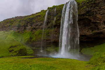 Front view of the waterfall. Water falls from rocks that are overgrown with green grass.
