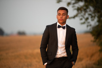 A handsome teenager dressed in black suit and bow tie and looks serious, He stands on a field that...