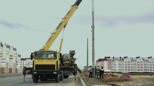 Workers install a pole with street light by automobile crane along the road. Construction machinery, concrete or cement mixer transport truck are on the work zone. Engineers set up lamppost on highway