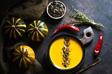 Homemade pumpkin soup with pumpkin seeds in a dark plate on a dark background with pumpkins, spices and herbs top view.