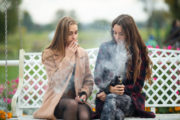 Smoking and vaping. Young beautiful white teeage girls in casual clothes sit on a vintage bench and smoke and vape cigarettes on the street in the evening. Electronic cigarette. Bad habit.