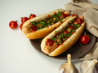 Hot dogi leżące na talerzu. Parówka, bułka, szczypiorek, pomidor. Jasne tło, widok z góry.