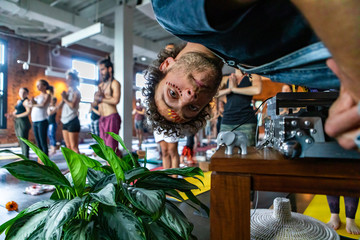 Diverse group of people in yoga class. A fun & playful Caucasian guy with painted face turns his head upside down and looks into the camera as blurred people attend a yogic session in the background.