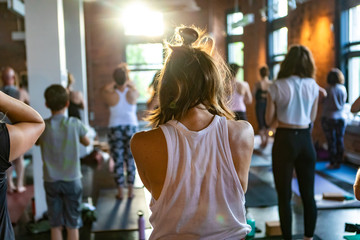 Diverse group of people in yoga class. A young Caucasian girl with blonde hair tied up is seen from behind during 108 rounds of surya namaskar (sun salutation). Blurred people are seen in background.