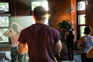 Diverse group of people in yoga class. A closeup and rear view of a healthy & muscular Caucasian man in a standing asana pose during 108 sun salutations, facing the sunlight during workout.