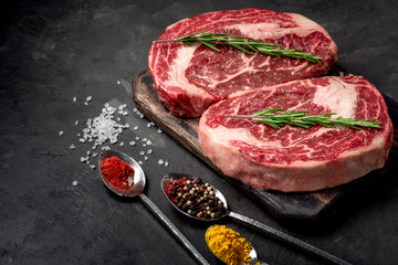 Two raw rib eye steak with seasonings on the dark stone background prepared for cooking. Marbled...