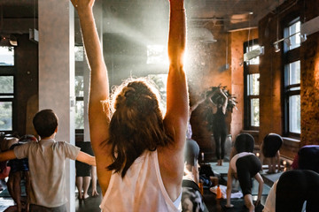 Diverse group of people in yoga class. A closeup and rear view of a young Caucasian woman with blonde hair and wearing a white top as she stands in upward hand stretch pose during 108 sun salutations