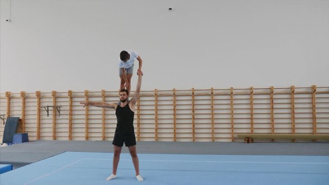 A young boy athlete is slowly making a handstand with one hand straight in side split on the head of a man partner in the sport center, lands on feet, steadicam, front view, side view