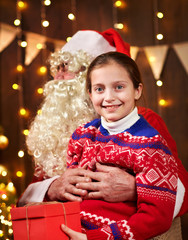 Santa Claus and child girl posing together indoor near decorated xmas tree with lights, they talking and smiling - Merry Christmas and Happy Holidays!