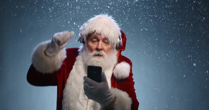 Funny Senior man in santa claus clothes and headphones is dancing to music played on his phone, isolated on blue snowy background - christmas spirit, christmas party close up 4k footage