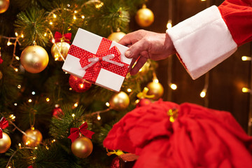 Closeup of Santa Claus hands with gift box, sitting indoor near decorated xmas tree with lights - Merry Christmas and Happy Holidays!
