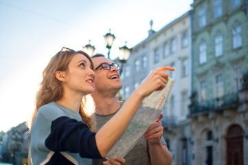Happy young tourists couple holding a paper map of ancient European city early in the morning on empty square and pointing on some sightseeing place