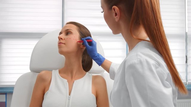 Beautician drawing correction lines on female patient's face