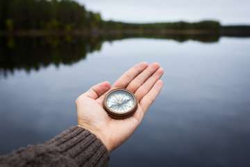Male hand holds a magnetic compass against the background of the forest and a lake. The concept of finding yourself the way and the truth.