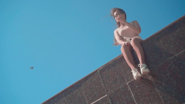 A girl in white shorts and a T-shirt with long hair walks along the city promenade. A girl sits on the granite fence of the embankment on the sky. Dutch angle