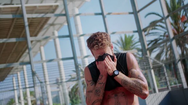 Portrait of tired sportsman after active training swiping sweat with shirt at hot sunny day, guy standing outdoor with metal fence on the background in great fit body shape watches on tattooed arms