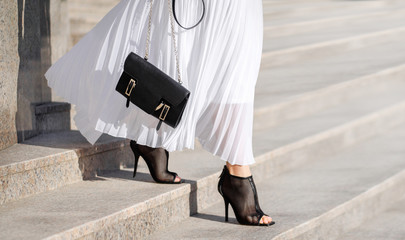 Fashionable bag close-up in female hands.Girl walks in the city outdoors. Stylish modern and feminine image, style. Girl in a white light dress with a black bag and black boots, or heeled shoes.