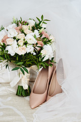 Bridal bouquet of pink, white roses and greenery with satin ribbon, beige women shoes and veil on bed, copy space. Wedding concept