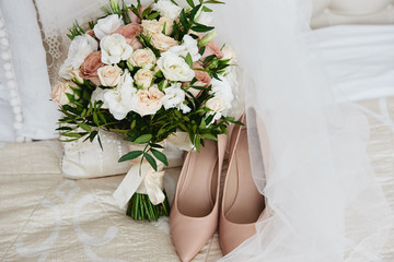 Bridal bouquet of pink, white roses and greenery with satin ribbon, beige women shoes and veil on bed, copy space. Wedding concept