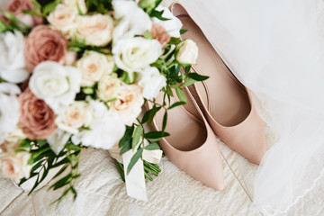 Bridal bouquet of pink, white roses and greenery with satin ribbon, beige women shoes and veil on bed, copy space. Wedding concept