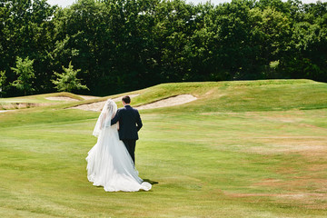 Full length body portrait of young bride and groom walking on green grass of golf course, back...