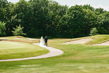 Full length body portrait of young bride and groom walking on green grass of golf course, back view. Happy wedding couple, copy space