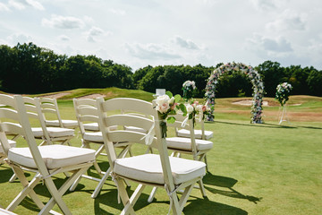 White wooden chairs with rose flowers on each side of archway outdoors, copy space. Empty chairs for guests prepared for wedding ceremony on golf course