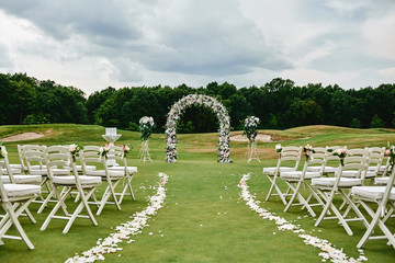 Place for wedding ceremony on green golf course, copy space. Wedding arch decorated with flowers and white chairs on each side of archway outdoors. Wedding setting