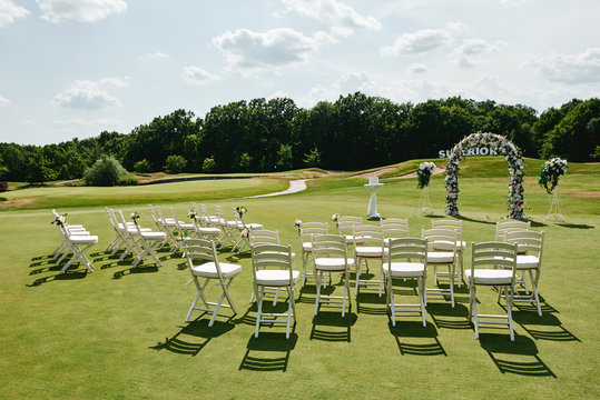 Place for wedding ceremony on green golf course, copy space. Wedding arch decorated with flowers and white chairs on each side of archway outdoors. Wedding setting