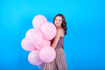 Beautiful young woman in dress having fun with pink helium air balloons over blue background. Holiday, birthday, valentine, fashion concept