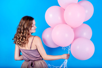 Beautiful young woman in dress holding pink balloons over blue background, back view. Holiday, birthday, valentine, fashion concept