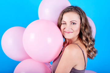 Cheerful young woman holding pink air balloons and wink isolated over blue background. Holiday, birthday, valentine, fashion concept