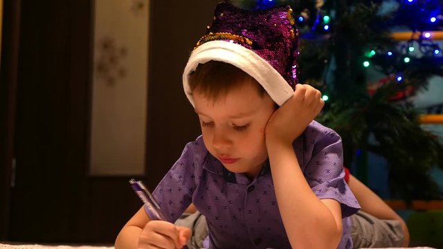 cute 4-6 years old boy writes a letter to Santa Claus in the background out of focus tree dressed up Santa Claus brings a gift