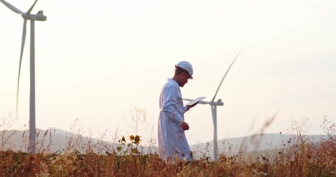 Technician in hard hat and uniform working with tablet computer modern technology at alternative wind power generating station.