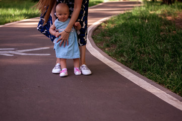 Mom teaches daughter to walk. Happy mom and daughter. Mom with a one-year-old daughter in dresses are walking in the park. Teaching children, motherhood. The first steps.