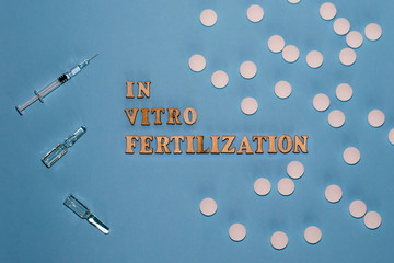 The inscription In Vitro Fertilization is in wooden letters on a blue background. In vitro injection pen, tablet and ampoule. A syringe with a thin huge. IVF concept. Infertility.