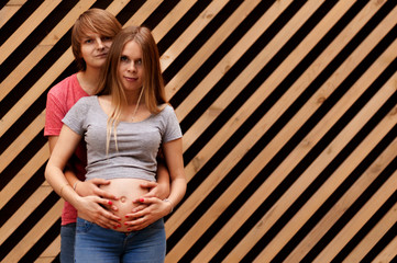 Happy young married couple, husband hugs a beautiful adult pregnant wife. Stand against the background of a striped wall. Second trimester Holds his hands on his stomach.