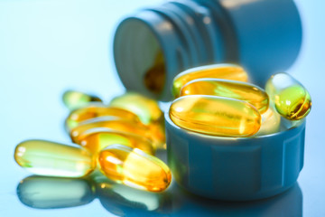 Omega 3 capsules spilled out from plastic bottle on blue gloss surface with reflections. Fish oil gold transparent capsules close-up, high resolution.