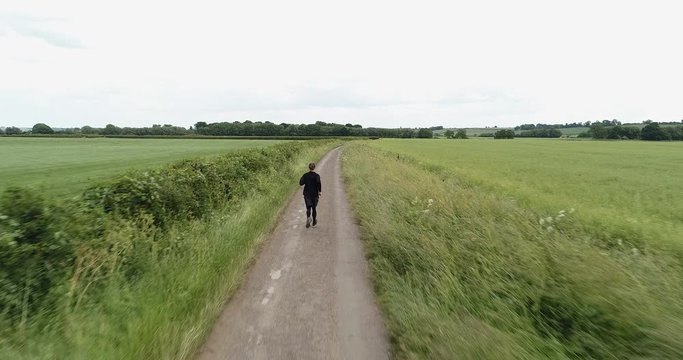 4K Drone of A Man Running on a Path  through Green Grass Fields. British  Countryside Farmers Land in the Summer. A Fitness Runner on a Long Dirt Road.