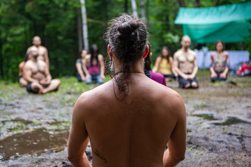 Diverse people enjoy spiritual gathering A slim topless guy with a hair bun is viewed from behind as people seek contemplation and enlightenment in natural surroundings.