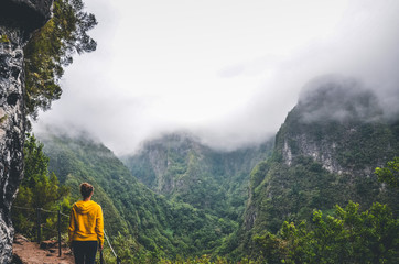 Young woman in yellow sweatshirt on a viewpoint in Levada Caldeirao Verde, Madeira, Portugal. Green scenic mountains in fog, misty landscape. Female traveler. Instagram, hipster filter - obrazy, fototapety, plakaty