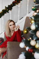 Blonde young woman in modish red dress posing near Christmas tree in the interior decorated for New Year