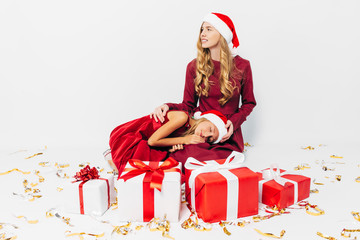 A young Christmas family, mom and daughter in Santa hats, spend time together sitting with...